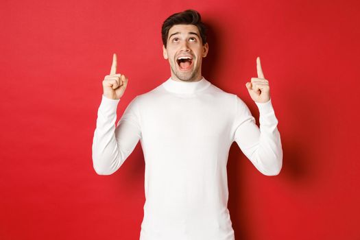 Concept of winter holidays. Image of amazed handsome guy reacting to christmas announcement, pointing and looking up at copy space, standing over red background.