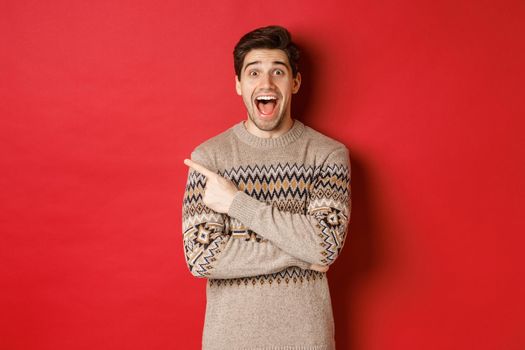 Portrait of amazed handsome guy, wearing christmas sweater, pointing finger at upper left corner logo and looking excited, showing awesome new year promo offer, standing over red background.