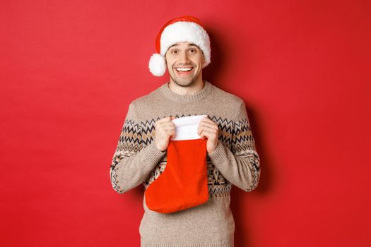 Concept of winter holidays, new year and celebration. Image of surprised and happy man receiving gift from secret santa inside christmas stocking bag, open gift and looking thankful.