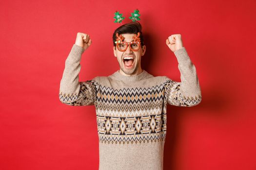 Portrait of thrilled and happy man in party glasses, winter sweater, shouting for joy and raising hands up, winning prize on christmas, achieve new year goal, standing red background.