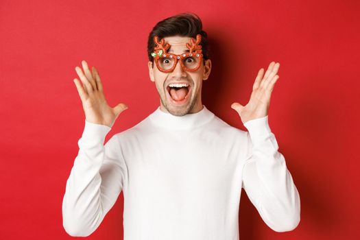 Close-up of surprised handsome guy in white sweater and party glasses, hear amazing christmas promo offer, rejoicing and standing over red background.