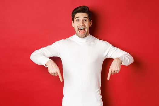 Image of amazed handsome guy in white sweater, reacting excited to christmas advertisement, pointing fingers down, showing logo, standing over red background.