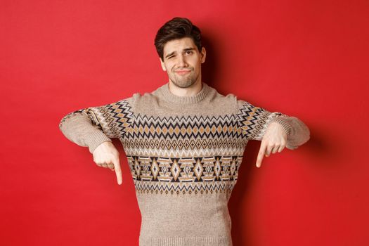 Concept of christmas celebration, winter holidays and lifestyle. Skeptical and unamused handsome man, showing something bad, pointing fingers down and grimacing, red background.