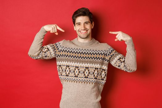 Concept of christmas celebration, winter holidays and lifestyle. Image of happy handsome man in xmas sweater pointing at himself and smiling, being secret santa, standing over red background.