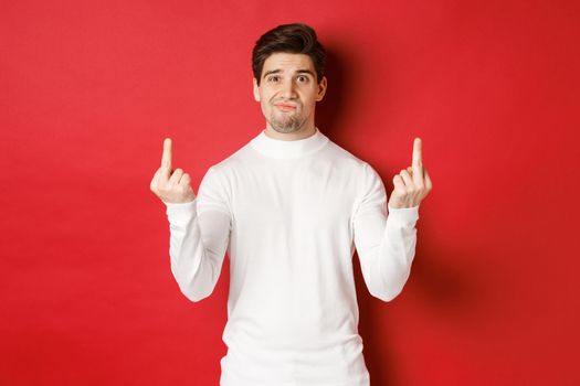 Image of pissed-off and distressed man telling to fuck off, showing middle-fingers and looking upset, standing over red background in white sweater.