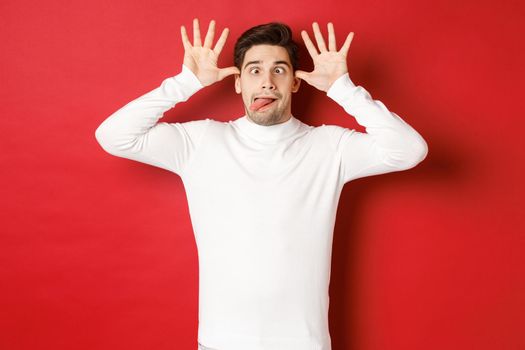 Portrait of funny caucasian guy, showing tongue and making faces, wearing white sweater, standing against red background.