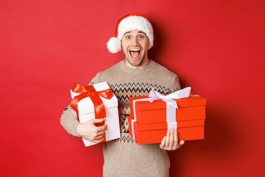 Concept of winter holidays, new year and celebration. Image of amazed and happy man in sweater and santa hat, holding christmas gifts and shouting for joy, red background.