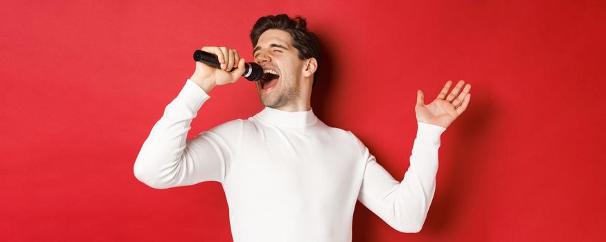 Handsome guy in white sweater, singing a song, holding microphone and performing at karaoke bar, standing over red background.