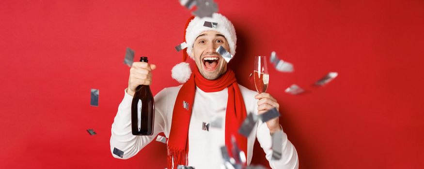 Concept of winter holidays, christmas and lifestyle. Close-up of happy man celebrating new year, holding champagne bottle and glass, standing over red background with confetti.