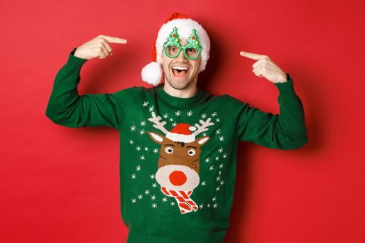 Image of excited smiling guy pointing at party glasses and celebrating new year, standing joyful in green sweater and santa hat against red background.