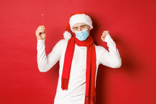 Concept of covid-19, christmas and holidays during pandemic. Happy man celebrating new year at party, wearing medical mask and santa hat, dancing with sparkler against red background.