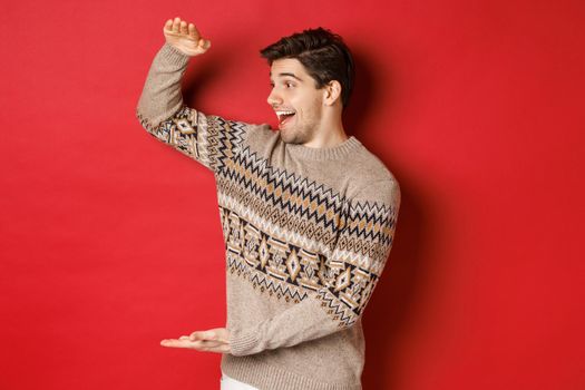 Portrait of happy young man in christmas sweater, showing large present, smiling and looking amazed at cool gift, standing over red background.