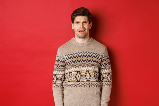 Portrait of confused adult man in christmas sweater, standing disappointed during new year holidays, dont understand something, standing over red background.