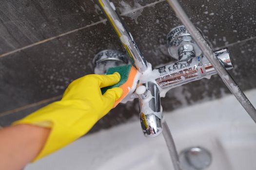 A hand in a yellow glove washes the mixer in the bathroom with a soapy sponge, close-up. Bathroom cleaning, disinfection