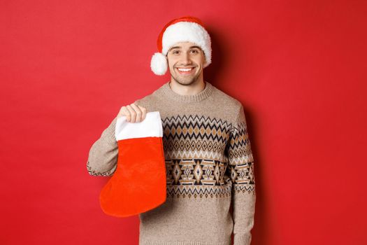 Concept of winter holidays, new year and celebration. Image of handsome smiling man in santa hat and sweater, holding christmas stocking for presents and candies, standing over red background.