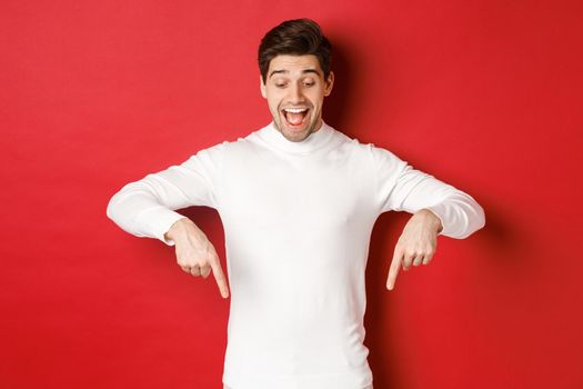 Portrait of happy good-looking man, pointing fingers down and looking at advertisement with excited smile, standing over red background.