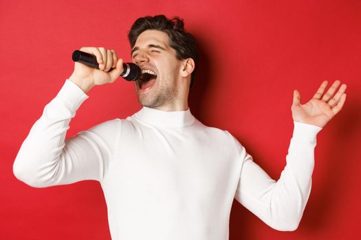 Handsome guy in white sweater, singing a song, holding microphone and performing at karaoke bar, standing over red background.