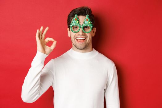 Happy caucasian guy wishing merry christmas, celebrating winter holidays, showing okay sign and smiling pleased, standing over red background.