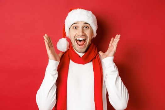 Concept of christmas, winter holidays and celebration. Image of handsome man looking surprised at new year promo offer, smiling amazed, standing over red background.