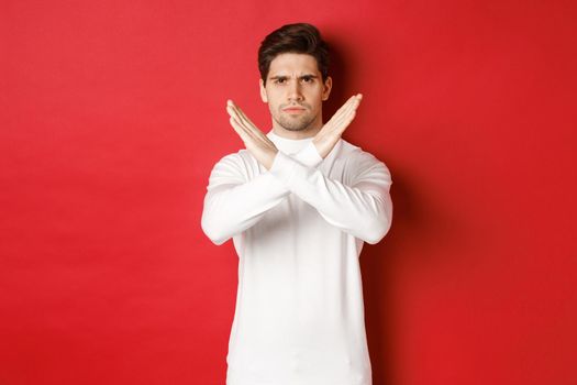 Image of serious and angry man in white sweater, express storng disapproval, showing cross sign to stop something bad, forbid action, standing over red background.