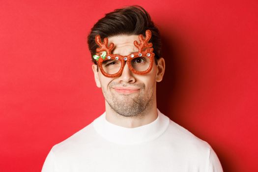 Concept of winter holidays, christmas and celebration. Close-up of skeptical handsome man in party glasses, looking doubtful and unamused, standing against red background.