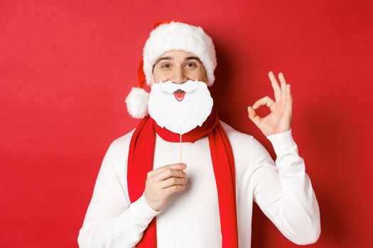 Concept of christmas, winter holidays and celebration. Pleased handsome man in santa hat, holding long white beard mask and showing okay sign, standing over red background.