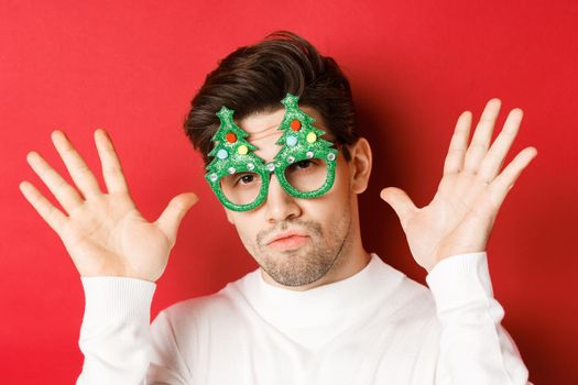Concept of winter holidays, christmas and celebration. Close-up of funny handsome guy in party glasses, showing empty hands, standing over red background.