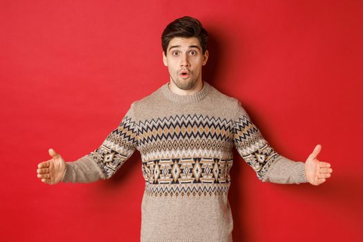 Image of amazed handsome man showing size of christmas present, spread hands sideways and shaping something large, standing over red background.