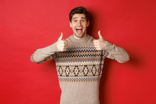 Portrait of cheerful man in christmas sweater like something good, showing thumbs-up and smiling amazed, praise new year party, standing over red background.