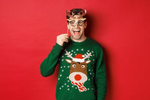 Handsome surprised man in christmas sweater, holding party mask and smiling, enjoying new year celebration, standing over red background.