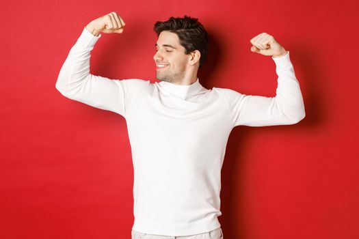 Portrait of smiling handsome man in white sweater, flexing biceps and bragging with strength, show-off strong muscles after workout, standing over red background.