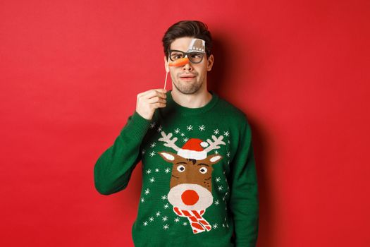 Funny man in christmas sweater and party mask, celebrating winter holidays, showing funny faces, standing over red background.