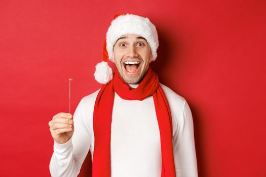 Concept of christmas, winter holidays and celebration. Close-up of happy handsome man in santa hat and scarf, having fun on new year party, holding sparkler and smiling, red background.