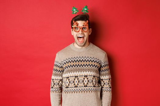 Image of surprised and excited handsome man, wearing party glasses with christmas sweater, drop jaw and looking at amazing new year promo offer, standing against red background.