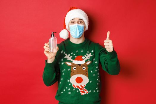 Concept of new year, coronavirus and social distancing. Portrait of cheerful man in santa hat, christmas sweater and medical mask, showing thumb-up and recommending hand sanitizer.