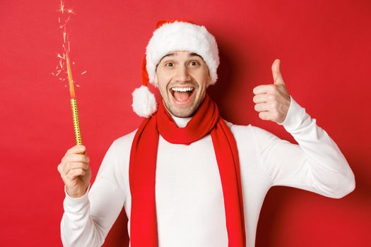Concept of christmas, winter holidays and celebration. Handsome man celebrating new year and having fun, holding sparkler and showing thumb-up, standing over red background.