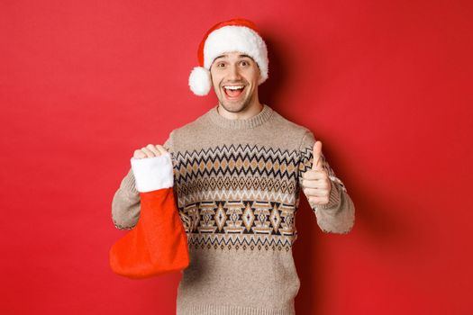 Concept of winter holidays, new year and celebration. Cheerful handsome man in santa hat and sweater, showing christmas stocking with candies and gifts, making thumbs-up.