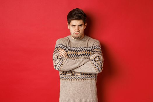 Image of angry and offedned guy in christmas sweater, feeling mad, cross arms on chest and sulking, standing over red background.