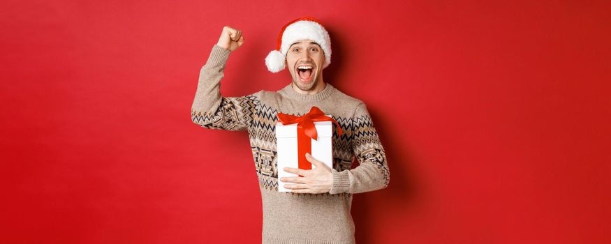 Image of happy and excited handsome man, reicing christmas gift, raising hands up in triumph and smiling, celebrating new year, standing over red background in santa hat.