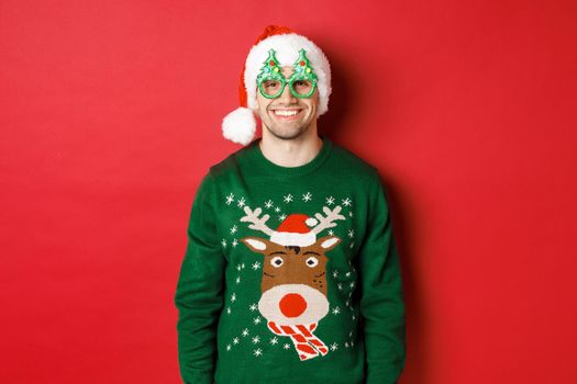 Portrait of handsome man in green christmas sweater, santa hat and party glasses, smiling happy, standing over red background.