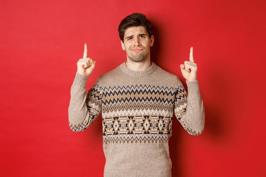 Image of skeptical and unamused handsome guy, showing bad christmas offer, pointing fingers up and grimacing displeased, express dislike, standing over red background.