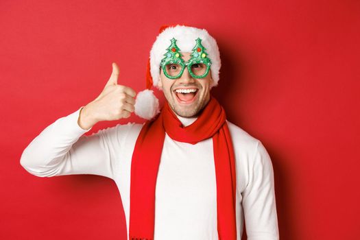 Concept of christmas, winter holidays and celebration. Cheerful guy in santa hat and party glasses, showing thumb-up and smiling pleased, standing over red background.