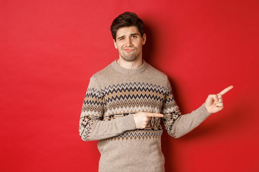 Concept of christmas celebration, winter holidays and lifestyle. Image of skeptical and displeased handsome man in xmas sweater, pointing fingers right and do not recommend product.