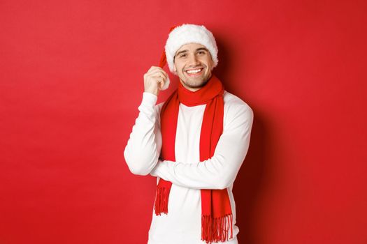 Portrait of happy man enjoying christmas holidays, wearing santa hat and scarf, smiling joyfully, standing over red background.