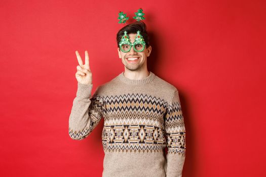 Portrait of funny and cute man in party glasses and christmas sweater, smiling and showing peace sign while posing over red background for new year advertisement.