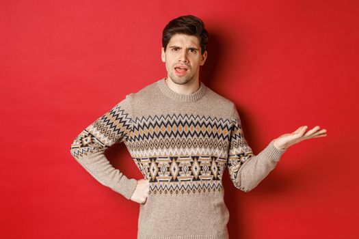 Image of disappointed and confused handsome man, wearing christmas sweater, raising hand and frowning, asking you to explain something, standing over red background.