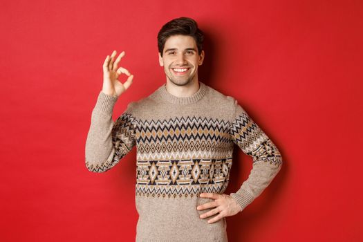 Concept of christmas celebration, winter holidays and lifestyle. Portrait of attractive man in xmas sweater, smiling happy and showing okay sign, praise something good, red background.