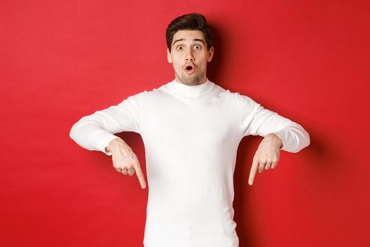 Image of surprised attractive guy in white sweater, showing announcement, pointing fingers down and looking amazed, standing over red background.