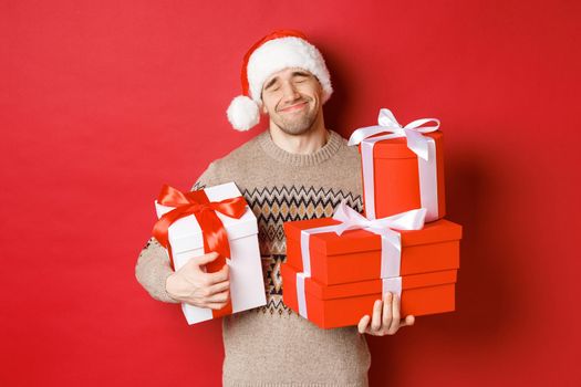 Concept of winter holidays, new year and celebration. Portrait of lovely smiling man receiving pile of presents, holding gifts and being touched with surprise, standing over red background grateful.