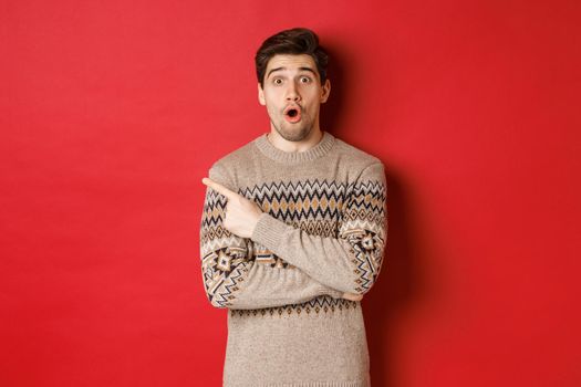 Image of impressed handsome guy, showing awesome christmas promo offer, pointing finger at upper left corner and looking amazed, standing in xmas sweater over red background.